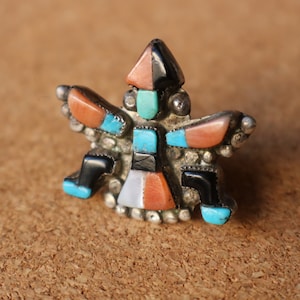 May include: A silver ring featuring a central figure inlay design composed of small, geometric pieces of turquoise, coral, black onyx, and white shell. The metal setting is decorated with small silver spheres along the outer edges of the figure.