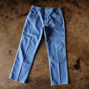 May include: Light blue denim pants with a classic five-pocket design. The pants have a straight leg cut and are made of a durable denim fabric. The pants are laid flat on a brown surface.
