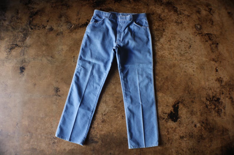 May include: Light blue denim pants with a classic five-pocket design. The pants have a straight leg cut and are made of a durable denim fabric. The pants are laid flat on a brown surface.