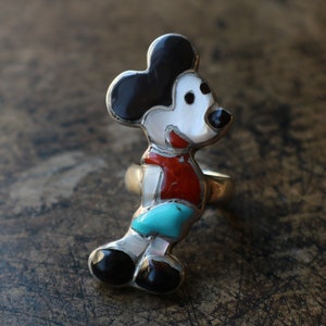 Peut inclure: Une bague en argent avec une incrustation de pierres multiples en forme de personnage de souris de dessin anim&eacute;. L'incrustation se compose de pierres noires, blanches, rouges et turquoise serties dans un cadre en argent poli.