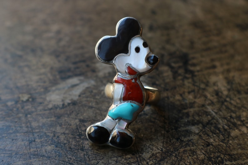 Peut inclure: Une bague en argent avec une incrustation de pierres multiples en forme de personnage de souris de dessin anim&eacute;. L'incrustation se compose de pierres noires, blanches, rouges et turquoise serties dans un cadre en argent poli.