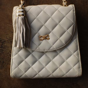 May include: A quilted, off-white handbag with a gold chain strap and a decorative tassel. The bag features a gold-colored clasp and a diamond pattern. The bag is approximately 8 inches wide and 9 inches tall.