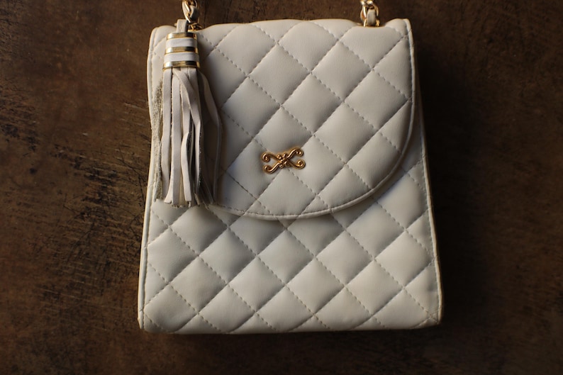May include: A quilted, off-white handbag with a gold chain strap and a decorative tassel. The bag features a gold-colored clasp and a diamond pattern. The bag is approximately 8 inches wide and 9 inches tall.