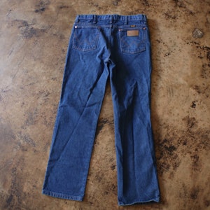 May include: A pair of dark blue denim jeans featuring a classic five-pocket design, a leather-like patch on the back right pocket, and signature W-shaped stitching on the rear pockets. The jeans have a button closure and belt loops.
