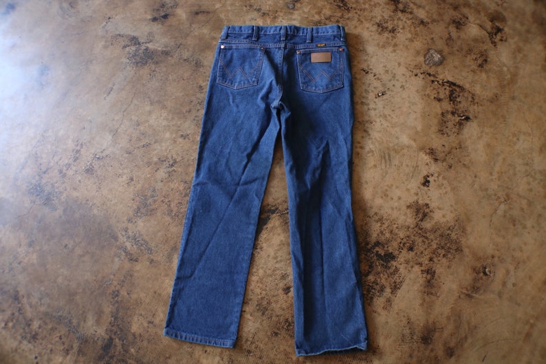 May include: A pair of dark blue denim jeans featuring a classic five-pocket design, a leather-like patch on the back right pocket, and signature W-shaped stitching on the rear pockets. The jeans have a button closure and belt loops.
