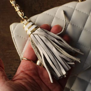 May include: White quilted handbag with a gold chain strap. The bag features a decorative tassel with gold accents. The bag is held by a hand, showcasing the details of the design. The bag's texture and color are prominent.