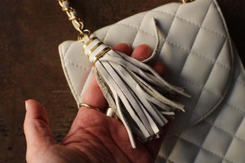 May include: White quilted handbag with a gold chain strap. The bag features a decorative tassel with gold accents. The bag is held by a hand, showcasing the details of the design. The bag's texture and color are prominent.