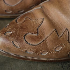 May include: Close-up of a pair of brown leather shoes. The shoes have decorative cut-out patterns on the toe and sides. The leather appears worn, with visible stitching and a textured surface. The shoes have a classic, vintage style.