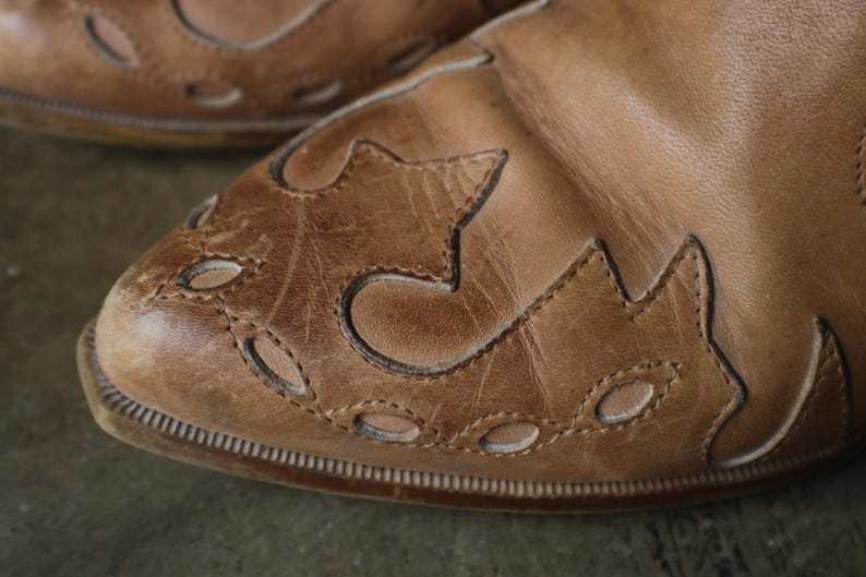 May include: Close-up of a pair of brown leather shoes. The shoes have decorative cut-out patterns on the toe and sides. The leather appears worn, with visible stitching and a textured surface. The shoes have a classic, vintage style.