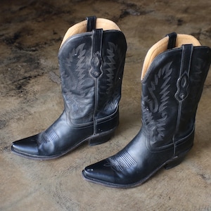 May include: A pair of black leather cowboy boots featuring a pointed toe, a stacked heel, and decorative tonal stitching on the shaft. The boots have a tan interior lining and pull tabs at the top of the shaft, showcasing a classic western design.