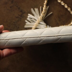May include: A white, quilted clutch with a gold chain strap and a tassel. The clutch has a diamond pattern and a zipper closure. The chain strap is made of small, interlocking links. The tassel is white and made of a soft material.