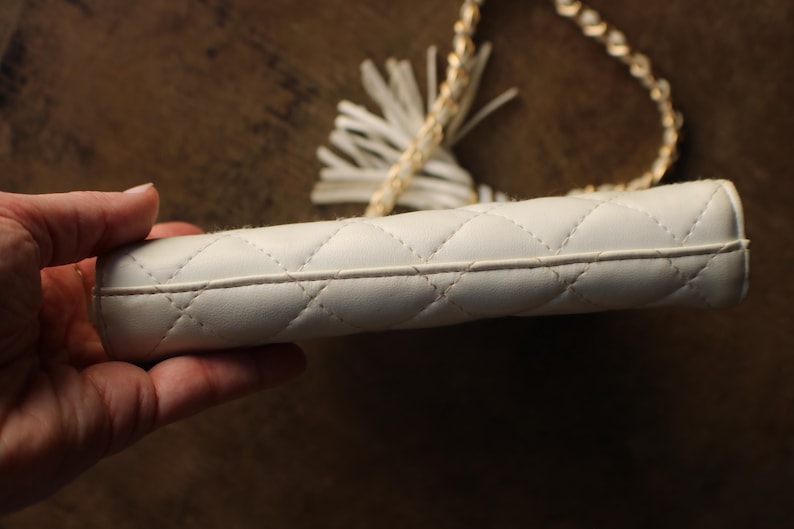May include: A white, quilted clutch with a gold chain strap and a tassel. The clutch has a diamond pattern and a zipper closure. The chain strap is made of small, interlocking links. The tassel is white and made of a soft material.