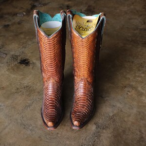 May include: A pair of brown cowboy boots featuring a textured snakeskin pattern on the exterior, pointed toes, and a turquoise interior lining. Each boot contains a yellow and white paper insert with the text CORRAL printed on the top.