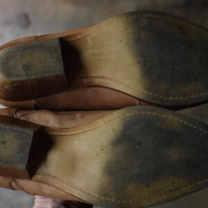 May include: A pair of worn, light brown leather cowboy boots, viewed from the soles. The soles show wear and tear, with a pointed toe and a stacked heel. The boots appear to be well-used.