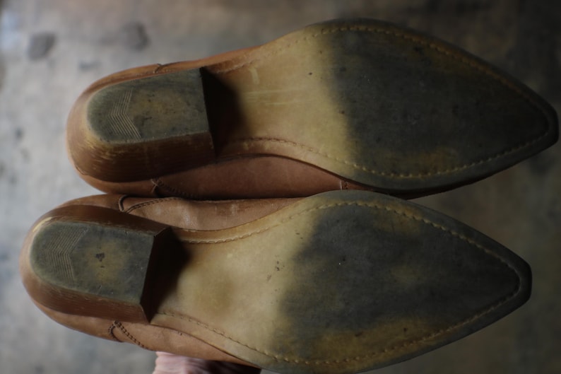 May include: A pair of worn, light brown leather cowboy boots, viewed from the soles. The soles show wear and tear, with a pointed toe and a stacked heel. The boots appear to be well-used.