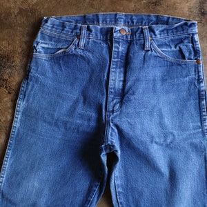 May include: Close-up of blue denim jeans. The jeans have a classic five-pocket design with a button and zipper closure. The denim has a slightly faded appearance, suggesting wear. The jeans are laid flat on a brown surface.