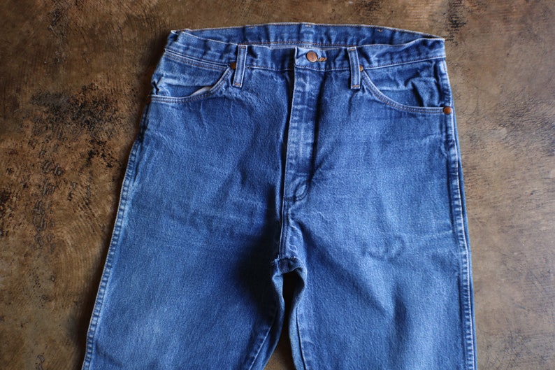 May include: Close-up of blue denim jeans. The jeans have a classic five-pocket design with a button and zipper closure. The denim has a slightly faded appearance, suggesting wear. The jeans are laid flat on a brown surface.