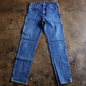 May include: Blue denim jeans with a classic five-pocket design. The jeans have a straight leg cut and a button and zipper closure. The denim has a faded wash, giving them a vintage look. The jeans are laid flat on a brown surface.