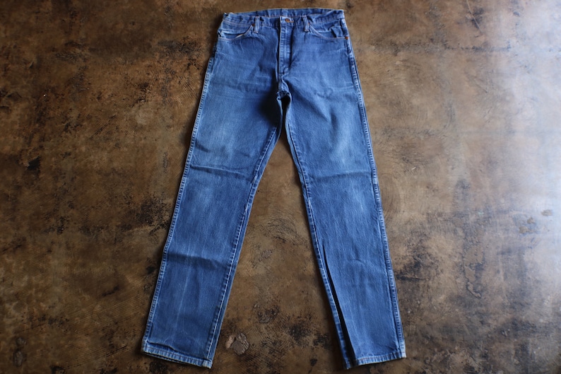 May include: Blue denim jeans with a classic five-pocket design. The jeans have a straight leg cut and a button and zipper closure. The denim has a faded wash, giving them a vintage look. The jeans are laid flat on a brown surface.