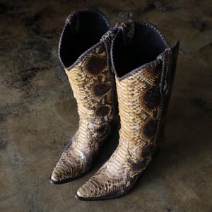 May include: A pair of knee-high cowboy boots featuring a textured snakeskin pattern in shades of tan, beige, and dark brown. The boots have a pointed toe, a tall shaft with a V-shaped opening at the top, and dark brown piping along the seams.