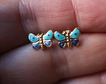 May include: A pair of gold-tone stud earrings shaped like butterflies. Each butterfly features light blue enamel on the upper wings with a small red dot, and dark blue glittery enamel on the lower wings, set against a polished gold metal body.