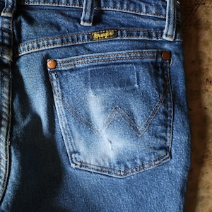 Puede incluir: Primer plano de la parte trasera de unos vaqueros azules. Los vaqueros tienen un bolsillo trasero con el logotipo de Wrangler y detalles de costura. El denim tiene un aspecto descolorido, con remaches de cobre y un aspecto desgastado.