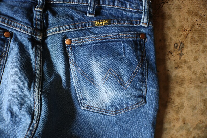 Puede incluir: Primer plano de la parte trasera de unos vaqueros azules. Los vaqueros tienen un bolsillo trasero con el logotipo de Wrangler y detalles de costura. El denim tiene un aspecto descolorido, con remaches de cobre y un aspecto desgastado.