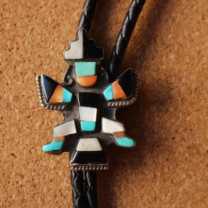 May include: A bolo tie featuring a central silver-toned metal ornament inlaid with geometric pieces of turquoise, coral, black onyx, and white mother-of-pearl. The ornament is attached to a braided black leather cord with a textured metal border.