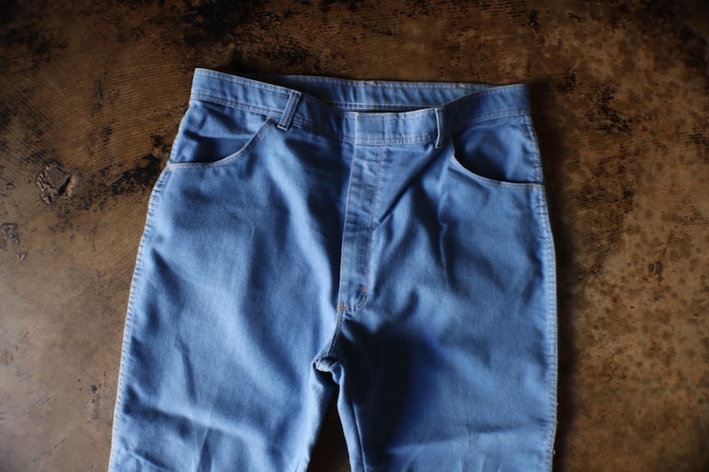 May include: Light blue denim pants with classic five-pocket design. The pants have a straight leg cut and a button and zipper closure. The fabric appears to be a medium-weight denim.
