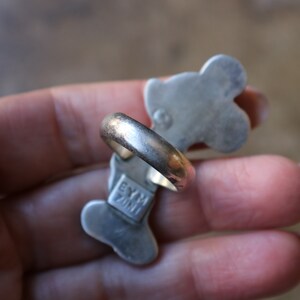 Peut inclure: Une bague en m&eacute;tal argent&eacute; pr&eacute;sentant une forme allong&eacute;e &agrave; plusieurs lobes. Le dessous de la bague affiche le texte estampill&eacute; EYM ZINI ainsi qu'un petit poin&ccedil;on circulaire. L'anneau est lisse et arrondi, avec une finition m&eacute;tallique.