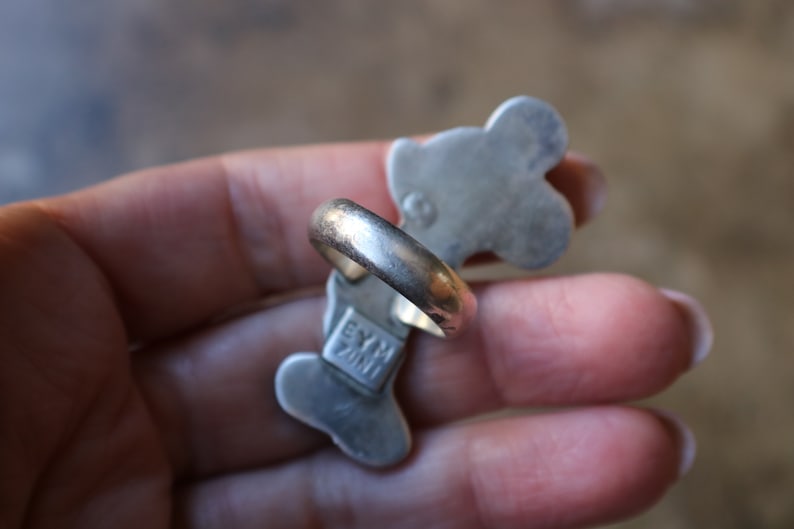 Peut inclure: Une bague en m&eacute;tal argent&eacute; pr&eacute;sentant une forme allong&eacute;e &agrave; plusieurs lobes. Le dessous de la bague affiche le texte estampill&eacute; EYM ZINI ainsi qu'un petit poin&ccedil;on circulaire. L'anneau est lisse et arrondi, avec une finition m&eacute;tallique.