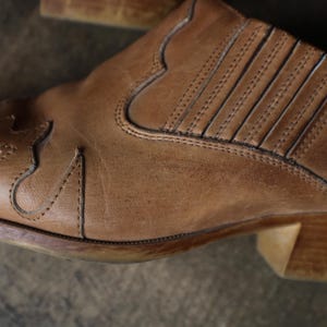 May include: Close-up of a brown leather boot. The boot features decorative stitching and a low heel. The leather has a worn appearance, with visible texture and subtle variations in color, suggesting age and use.