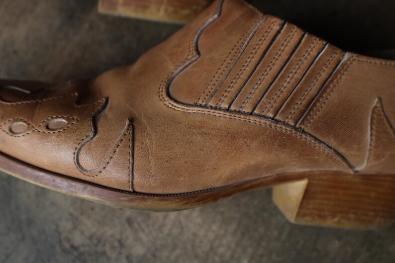 May include: Close-up of a brown leather boot. The boot features decorative stitching and a low heel. The leather has a worn appearance, with visible texture and subtle variations in color, suggesting age and use.