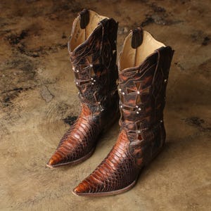 May include: A pair of dark brown leather cowboy boots featuring a pointed toe, a textured snakeskin-patterned vamp, and a tall shaft decorated with cut-out cross shapes and small silver-toned metal studs along the upper edges and shaft.
