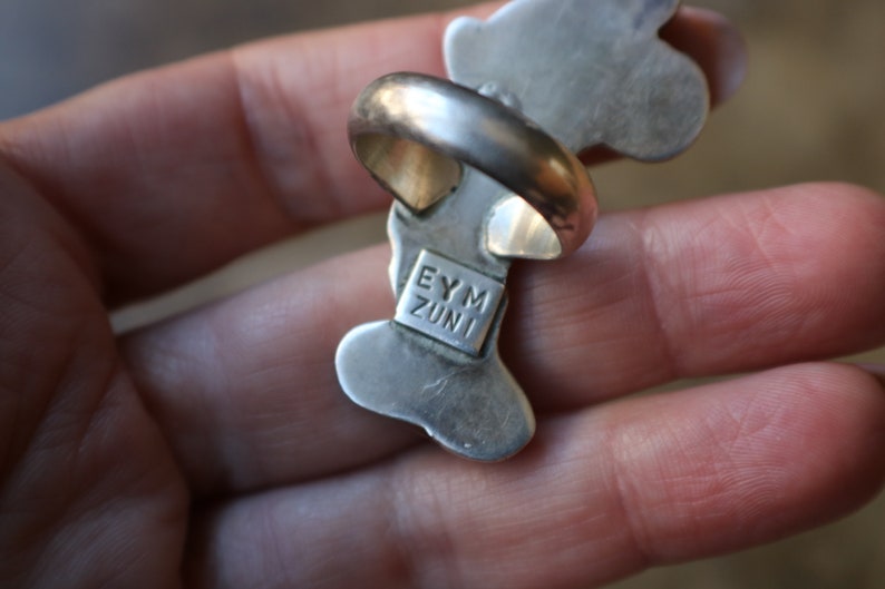 Peut inclure: Une bague en argent dot&eacute;e d'un anneau lisse et arrondi ainsi que d'une monture allong&eacute;e aux formes organiques. Le dessous de la bague pr&eacute;sente une plaque de poin&ccedil;on rectangulaire grav&eacute;e du texte EYM ZUNI en lettres capitales, centr&eacute;e.