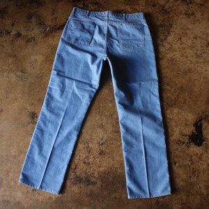 May include: Light blue denim pants with two back pockets and a classic straight leg design. The pants are laid flat on a brown surface, showcasing the back view of the garment. A casual style.