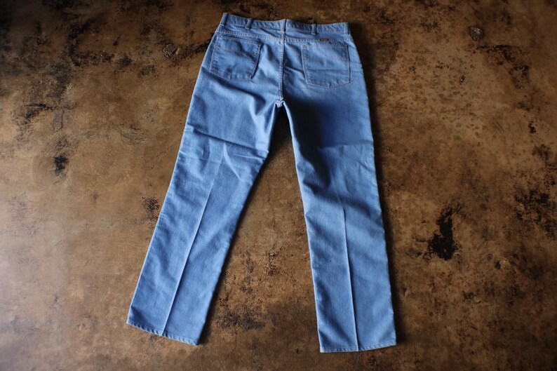 May include: Light blue denim pants with two back pockets and a classic straight leg design. The pants are laid flat on a brown surface, showcasing the back view of the garment. A casual style.