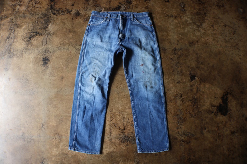 Puede incluir: Vaqueros de mezclilla azul descolorido con un corte recto cl&aacute;sico. Los vaqueros muestran signos de desgaste, con manchas y decoloraci&oacute;n visibles, lo que les da una est&eacute;tica vintage. Los vaqueros est&aacute;n colocados sobre una superficie marr&oacute;n.
