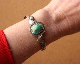 May include: Silver cuff bracelet featuring a central turquoise stone. The bracelet has a detailed, leaf-like design on the sides of the turquoise stone and along the band. The turquoise stone is a vibrant green-blue color.