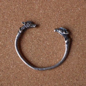 May include: A silver-toned open cuff bracelet. The bracelet features a circular design with animal head details at each open end. The bracelet is set against a brown, textured background.