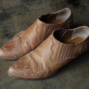 May include: A pair of brown leather slip-on shoes. The shoes have a pointed toe and decorative cut-out patterns on the front. The sides feature elastic panels for easy wear. The shoes are displayed on a textured surface.