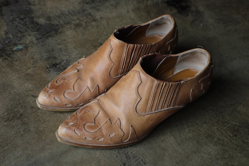 May include: A pair of brown leather slip-on shoes. The shoes have a pointed toe and decorative cut-out patterns on the front. The sides feature elastic panels for easy wear. The shoes are displayed on a textured surface.