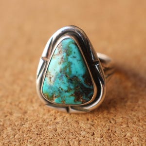 May include: A silver ring featuring a triangular turquoise stone with dark brown matrix patterns. The metal setting has a smooth, polished finish with decorative notched details along the outer edge surrounding the central gemstone.