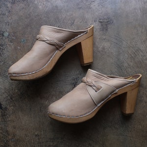 May include: A pair of beige leather clogs featuring a braided strap detail across the vamp, a rounded toe, and a sturdy wooden block heel. The leather upper is attached to the wooden sole with visible silver-toned metal staples along the perimeter.