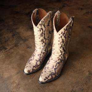 May include: A pair of cowboy boots made from snakeskin. The boots have a beige and brown pattern, with a pointed toe and a tall shaft. The boots are set against a brown background.