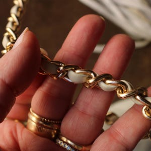 May include: Close-up of a gold chain strap with white leather woven through the links. The chain is held in a hand with multiple gold rings on the fingers. The background is blurred.