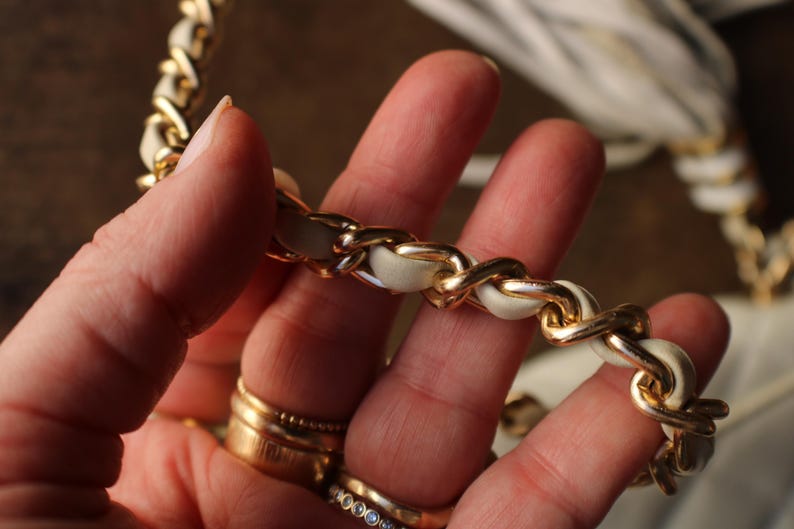 May include: Close-up of a gold chain strap with white leather woven through the links. The chain is held in a hand with multiple gold rings on the fingers. The background is blurred.