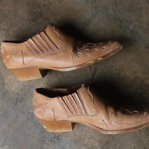 May include: A pair of tan leather ankle boots with a Western-inspired design. The boots feature decorative stitching and a low heel. The leather appears worn, giving them a vintage aesthetic. The boots are set against a neutral background.