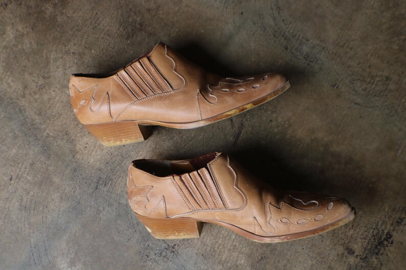 May include: A pair of tan leather ankle boots with a Western-inspired design. The boots feature decorative stitching and a low heel. The leather appears worn, giving them a vintage aesthetic. The boots are set against a neutral background.