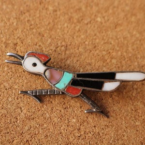 May include: A roadrunner-shaped brooch crafted from silver metal with inlaid stone segments. The bird features a white head, red crest, pink and turquoise chest, and black and white tail feathers, all set within a thin silver frame.